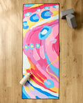 The "Abundance" recycled yoga mat towel by Sareka Unique, featuring vibrant pink, blue, yellow, and white abstract patterns, rests on a wooden floor beside two gray yoga blocks and a white water bottle with a pink lid.