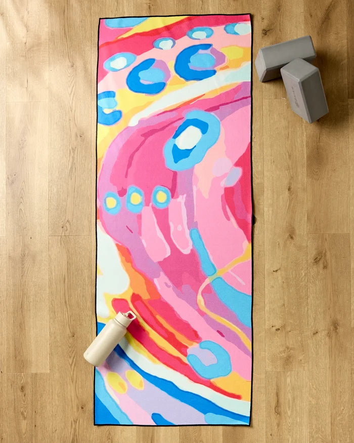 The "Abundance" recycled yoga mat towel by Sareka Unique, featuring vibrant pink, blue, yellow, and white abstract patterns, rests on a wooden floor beside two gray yoga blocks and a white water bottle with a pink lid.