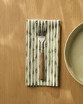 A Bamboo fork with a wooden handle rests on a Geometry Geosuede recycled dinner napkin beside a light green plate, all on a light wooden table.