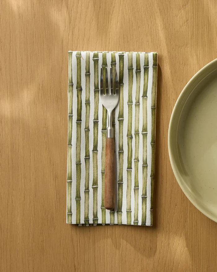 A Bamboo fork with a wooden handle rests on a Geometry Geosuede recycled dinner napkin beside a light green plate, all on a light wooden table.