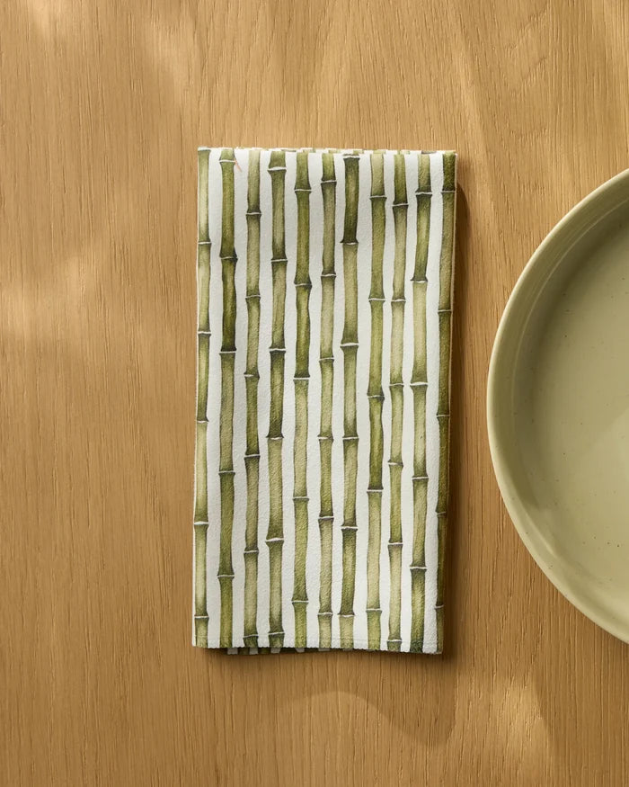 A Geometry "Bamboo" recycled dinner napkin with a green bamboo pattern sits folded on a light wooden table beside a round pale green plate.