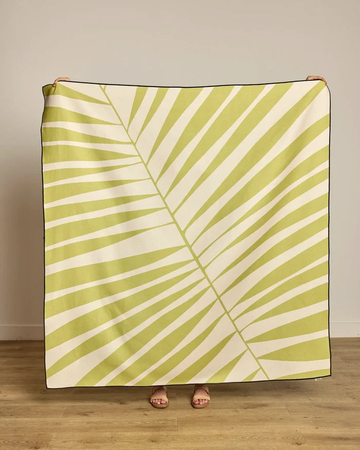 A person stands behind the Geometry Cabana Club beach blanket, featuring a light green palm leaf design on cream. Made from recycled materials, only their hands and feet show against a plain wall and wooden floor.