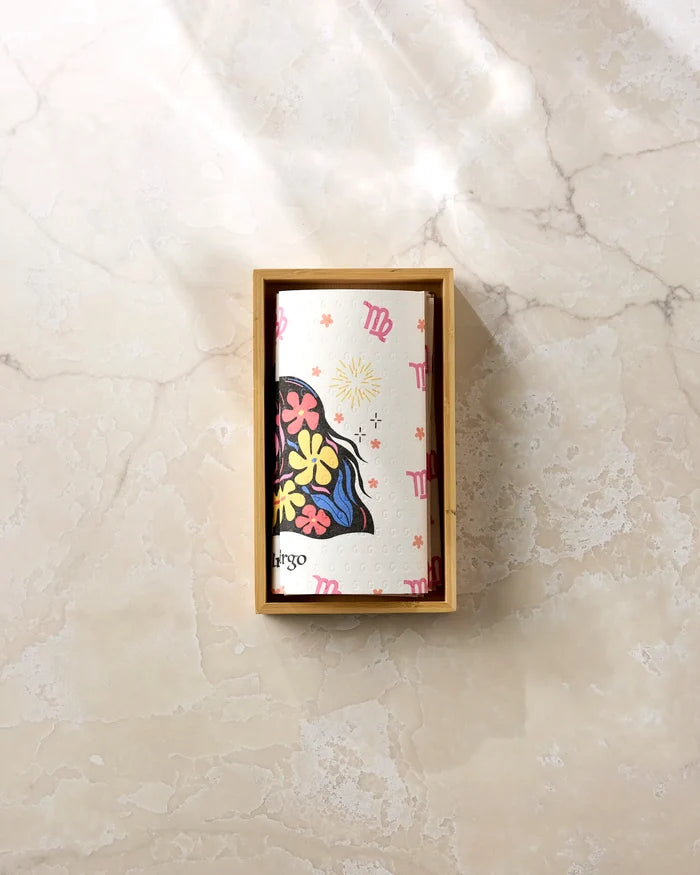 The Clever Virgo card by Heather Zhou, featuring floral patterns and Virgo symbols, sits in a small wooden box on light marble next to eco-friendly kitchen towels. Sunlight casts gentle shadows across the stylish arrangement.