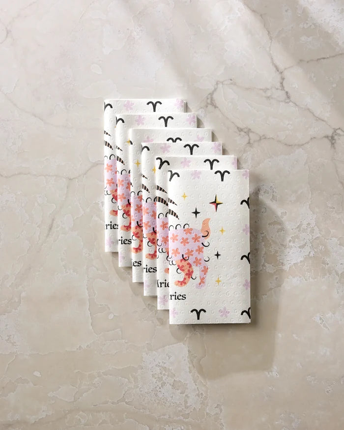 Six folded "Daring Aries" cloth napkins by Heather Zhou, featuring ram horns, stars, and "Aries," are arranged diagonally on marble—a stylish, eco-friendly alternative to paper towels for your home.