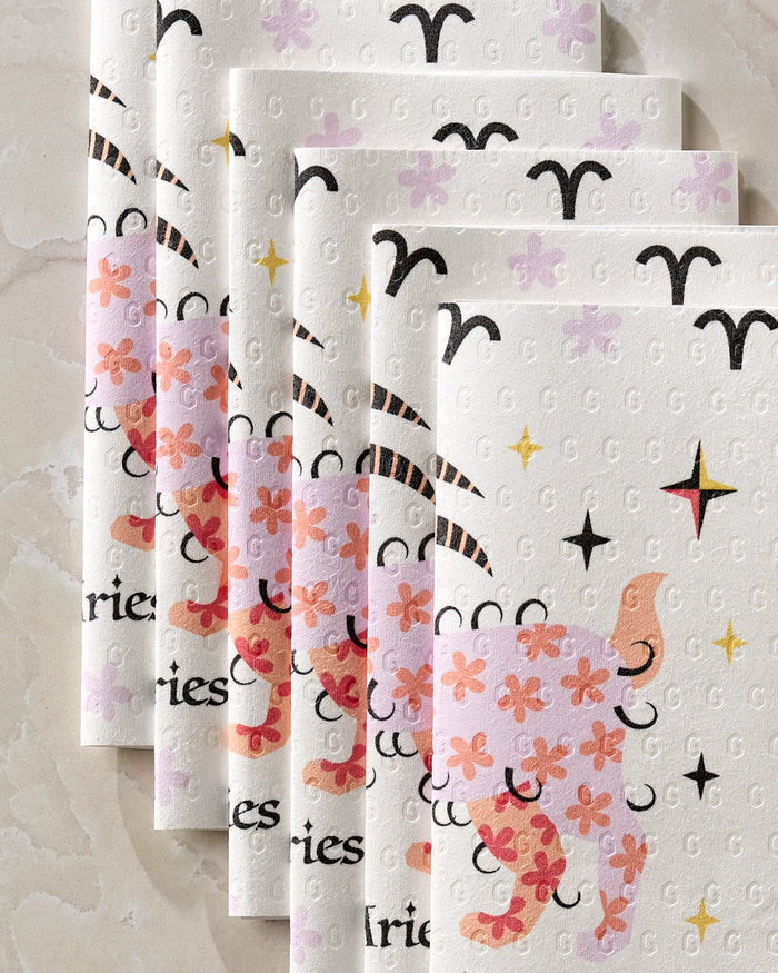 Six sheets of Heather Zhou's Daring Aries eco-friendly paper towels, featuring pink and orange cartoon rams with black Aries zodiac symbols, are arranged in a staggered stack on a light marble surface.