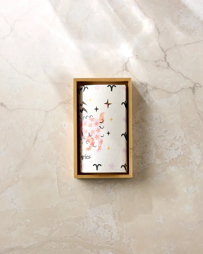 The "Daring Aries" card by Heather Zhou, featuring zodiac art and a ram, rests partly inside a wooden box on a marble surface beside eco-friendly paper towels in gentle sunlight.
