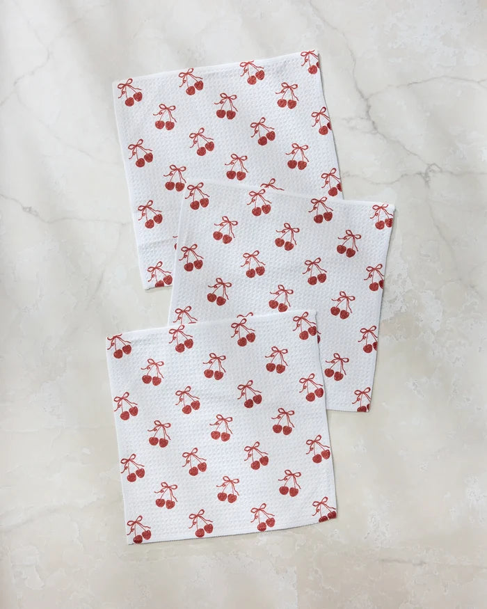 Three "Darling" napkins by Geometry, made from recycled materials and featuring a repeating pattern of red cherries and small bows, are arranged on a light marble surface.