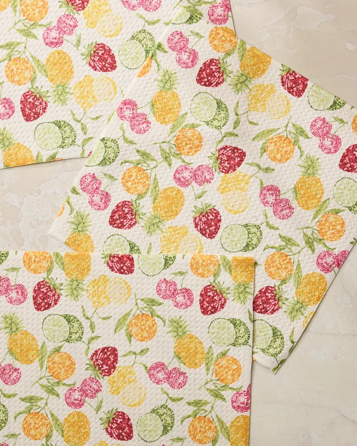 Three white Disco Fruits dishcloths by Geometry, featuring colorful prints of pineapples, limes, lemons, strawberries, and cherries, are displayed on a light beige textured surface.