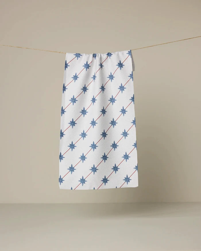 The Estrella Norte towel by Geometry, featuring blue eight-pointed stars in diagonal lines on a white background, hangs on a clothesline against plain beige.