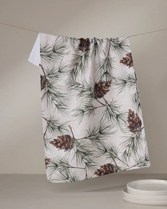 The Geometry "Evergreen Frost" kitchen towel, featuring a pinecone and pine needle pattern, hangs from a clothesline above a stack of white dishes on a light-colored surface.