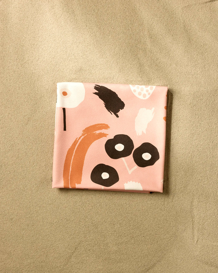 The folded Fall Flowerbed beach blanket by Marina Solodka features pink, abstract black, white, and brown shapes on a beige surface. Made from quick-drying recycled materials, it’s ideal for eco-conscious days by the shore.
