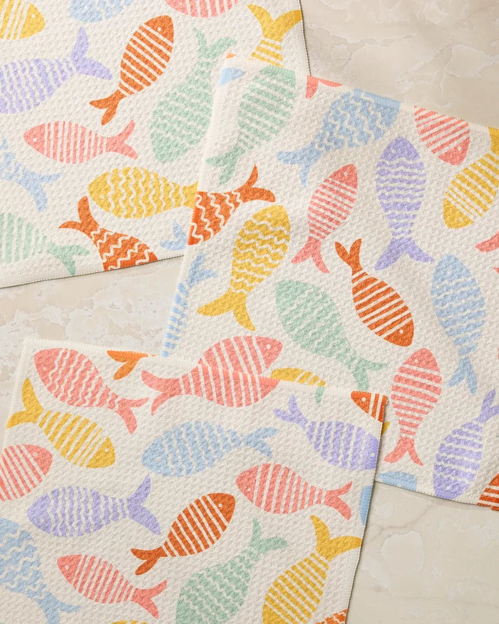 Three Fish Flip by Geometry dishtowels, each super-absorbent and featuring unique colorful fish in orange, yellow, green, pink, and purple with stripes or zigzags, lie slightly overlapping on a light beige surface.