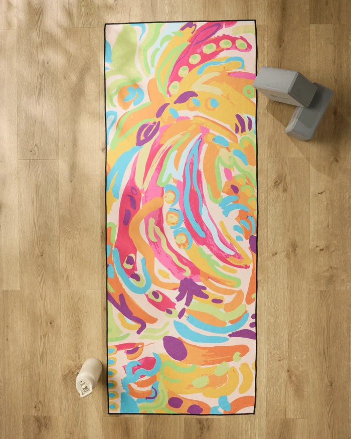 The Florescence yoga mat by Sareka Unique, featuring vibrant abstract patterns in orange, pink, blue, green, and purple, is on a wooden floor alongside a white water bottle, two gray yoga blocks, and a silicone grip yoga towel.