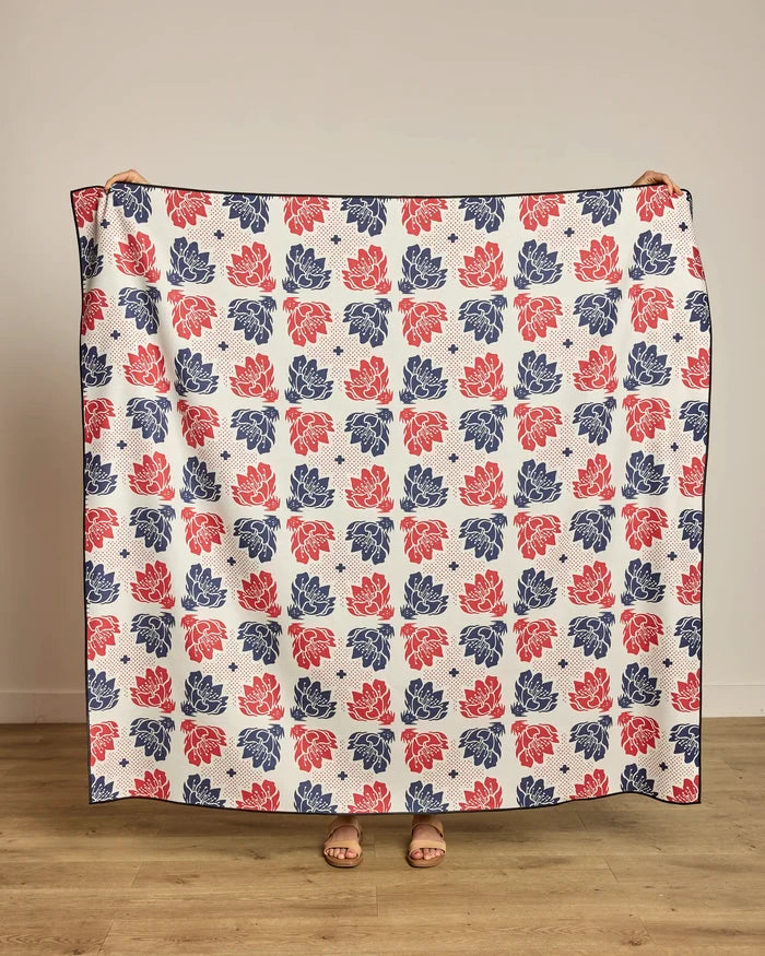 A person stands on a wooden floor, showing only their hands and feet as they hold up Geometry’s Founding Floral beach blanket—a large quilt-style piece made from recycled materials, featuring a red and navy floral grid on white.