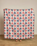 A person stands on a wooden floor, showing only their hands and feet as they hold up Geometry’s Founding Floral beach blanket—a large quilt-style piece made from recycled materials, featuring a red and navy floral grid on white.