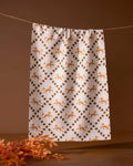 The Geometry Foxes and Twinkles tea towel, featuring an orange and brown floral and leaf design, hangs on a clothesline against a brown background with orange foliage in the bottom left.