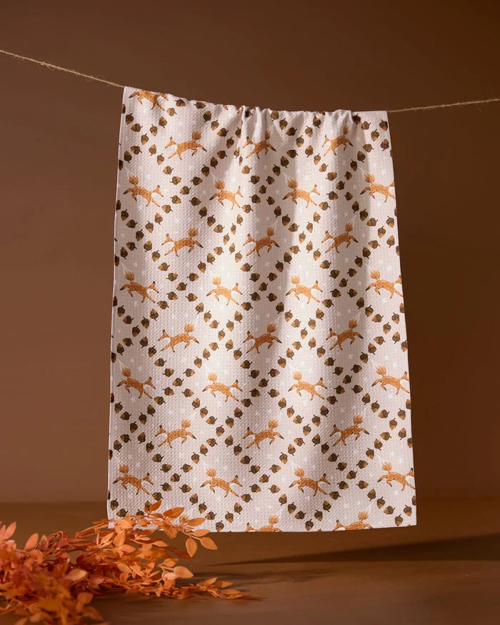 The Geometry Foxes and Twinkles tea towel, featuring an orange and brown floral and leaf design, hangs on a clothesline against a brown background with orange foliage in the bottom left.