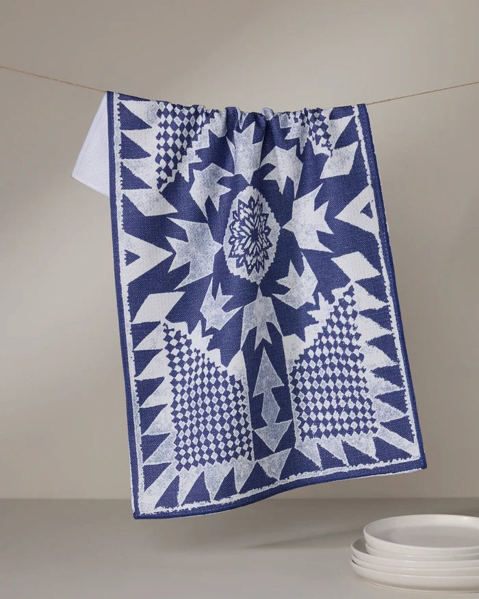 The Geometry Froststar kitchen towel, featuring blue and white geometric star patterns, hangs above a stack of white plates against a neutral background.