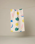 The Geometry Fruity Pop kitchen towel, featuring a vibrant print of watermelon slices, ice cream popsicles, flowers, cherries, and monstera leaves on beige fabric, hangs on a clothesline against a plain backdrop.