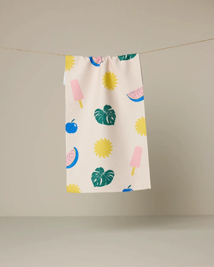 The Geometry Fruity Pop kitchen towel, featuring a vibrant print of watermelon slices, ice cream popsicles, flowers, cherries, and monstera leaves on beige fabric, hangs on a clothesline against a plain backdrop.