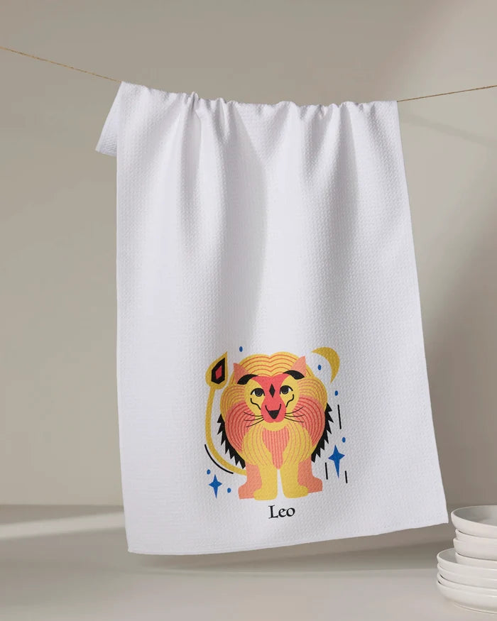 The Glowing Leo tea towel by Heather Zhou hangs on a line, displaying a vibrant cartoon lion and "Leo" below. Made from quick-drying recycled materials, it brightens your kitchen. A stack of white plates appears in the lower right corner.