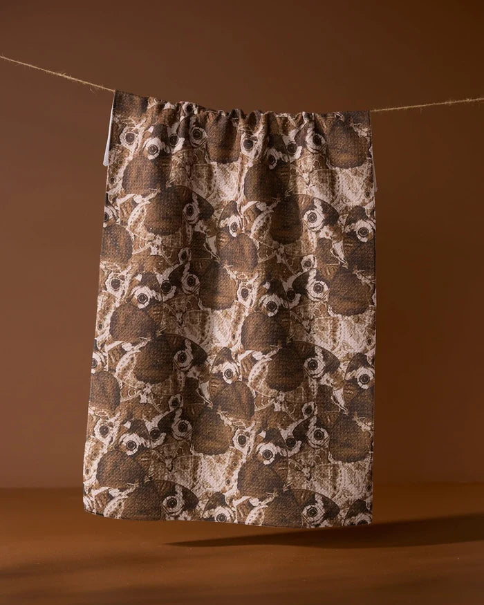The Geometry Gossamer Flight tea towel, made from soft Geoweave fabric, features an illustrated pattern of owl faces and hangs on a string against a solid brown background.