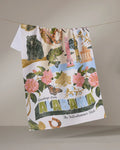 The Geometry "Greetings From Alabama" tea towel, ultra-absorbent and colorful, hangs on a line featuring Alabama symbols—flowers, animals, landmarks—and the text “Greetings from ALABAMA The Yellowhammer State.”.