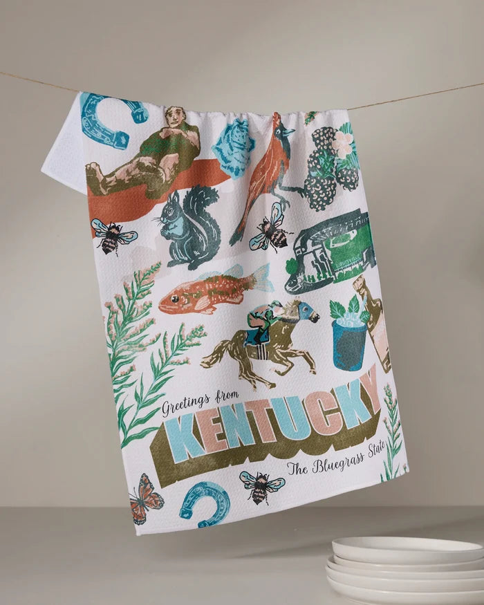 A recycled Geometry kitchen towel, “Greetings From Kentucky,” hangs on a line, featuring colorful prints of a horse, jockey, fish, flowers, bees, horseshoes, and “Greetings from KENTUCKY The Bluegrass State.”.