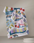 The Geometry "Greetings From Michigan" tea towel features Michigan icons—turtles, fish, cherries, flowers, the Mackinac Bridge, and “Greetings from Michigan: The Great Lakes State”—and hangs near white plates. Ultra-absorbent and colorful.