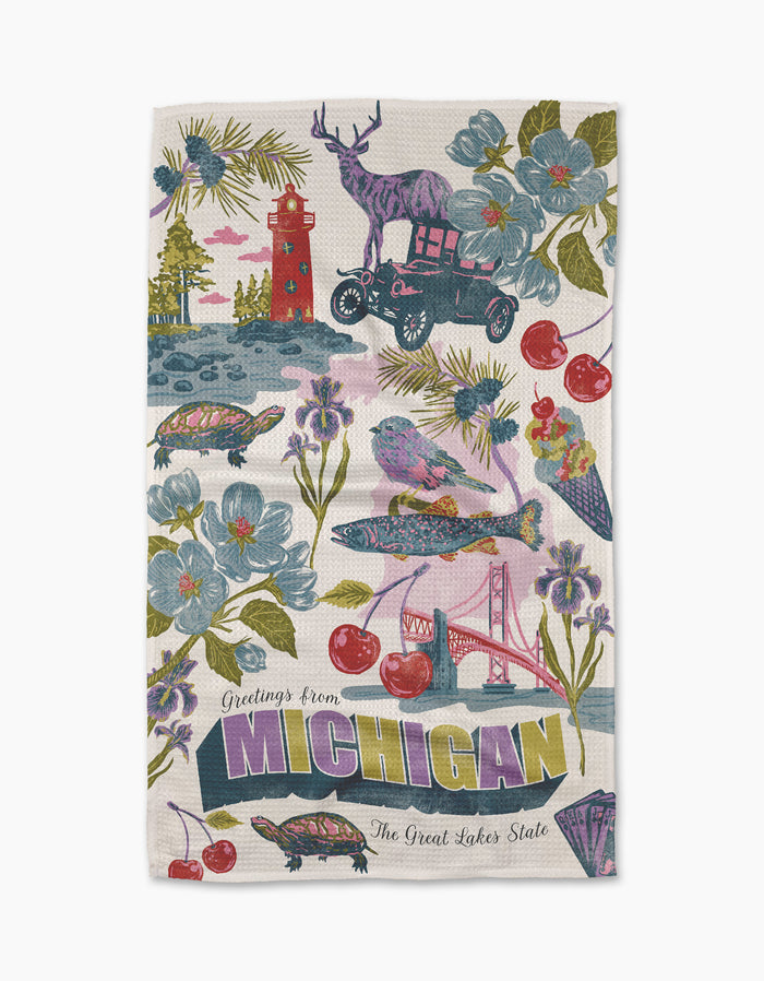 The Geometry "Greetings From Michigan" tea towel features colorful Michigan symbols—deer on a car, lighthouse, cherries, turtles, birds, flowers, sailboat, and the Mackinac Bridge—with "Greetings from Michigan The Great Lakes State" text.