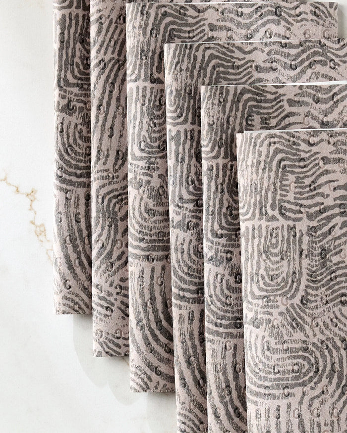 Five “Imprint” cloth napkins by Studio Jeannot, featuring a gray and black abstract fingerprint pattern, are elegantly stacked on a white marble surface with subtle gold veining—a chic alternative to disposable paper towels.