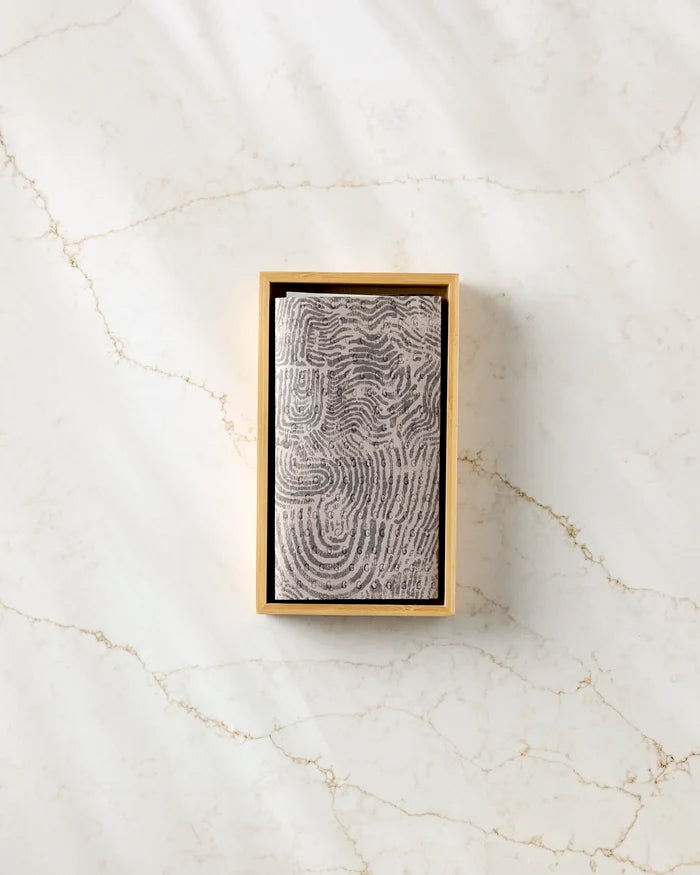 The Imprint reusable gray paper towel by Studio Jeannot, featuring a black fingerprint pattern, is folded inside a rectangular beige box on a white marble surface with light brown veining.