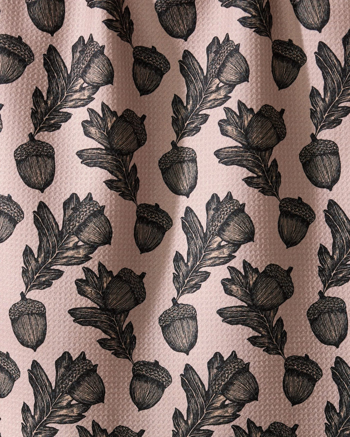 Geometry’s Kindred dish towel features an illustrated pattern of black acorns and oak leaves, evenly spaced on a textured light pink background. Ultra-absorbent fabric forms soft folds with subtle shadows and highlights.