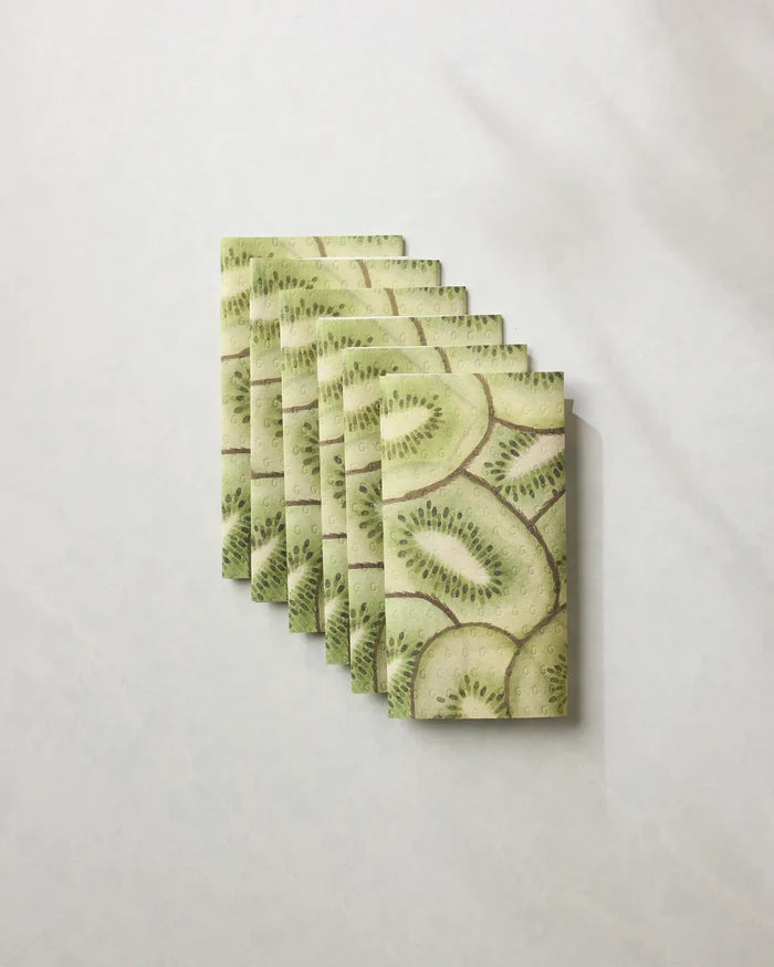 Nine rectangular crackers topped with Geometry Kiwi Slices are arranged in an overlapping row on a surface resembling eco-friendly kitchen towels, with shadows to the right enhancing the minimalist look.