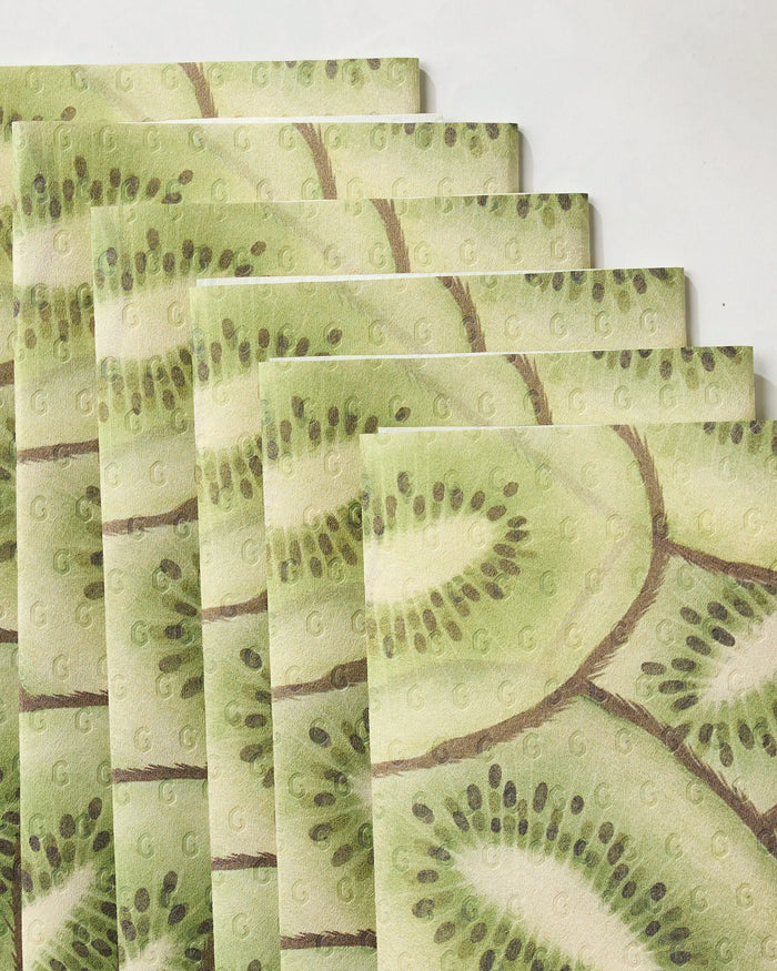 Six folded Geometry Kiwi Slices eco-friendly kitchen towels are arranged in a staggered stack. Each towel features a vibrant kiwi slice pattern against a pale green background.