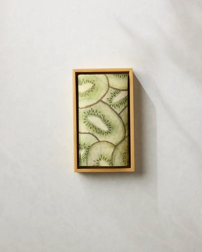 A rectangular wooden tray holds Geometry’s Kiwi Slices dessert, topped with neatly layered fresh kiwi, displayed on a light textured surface next to folded reusable paper towels.