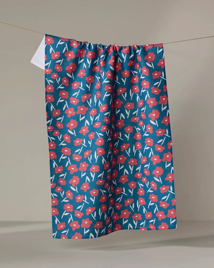 The Hufton Studio Luca Daisy Red tea towel, featuring a red and white floral design on blue, is displayed hanging against a plain beige background.