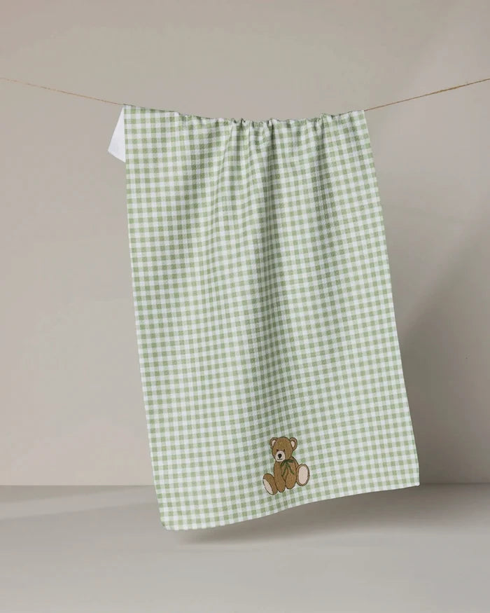 The Geometry Lucky Bear kitchen towel features green and white gingham with an embroidered brown teddy bear, shown hanging on a clothesline against a beige background.