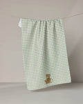 The Geometry Lucky Bear kitchen towel features green and white gingham with an embroidered brown teddy bear, shown hanging on a clothesline against a beige background.