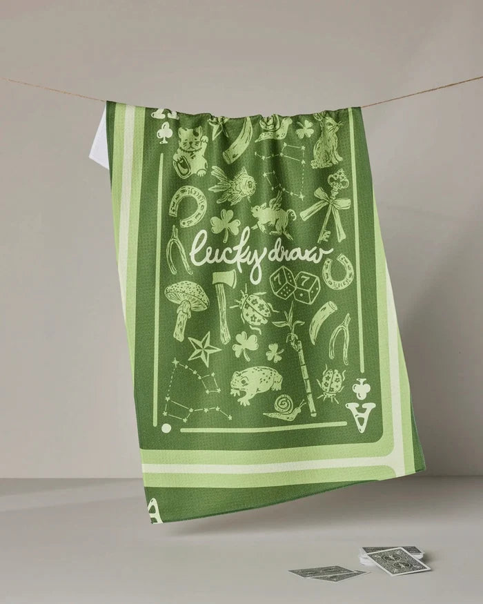 The Geometry Lucky Draw green absorbent dish towel, featuring "lucky draw" in white and decorated with luck-themed icons like horseshoes, clovers, dice, and a lucky cat, hangs over a clothesline.