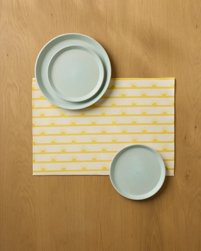 Two light blue plates rest on a wooden table, set atop the Geometry Luminary Placemat, which features a repeating yellow and white sun pattern. The larger plate is positioned at the top left, while the smaller is at the bottom right.