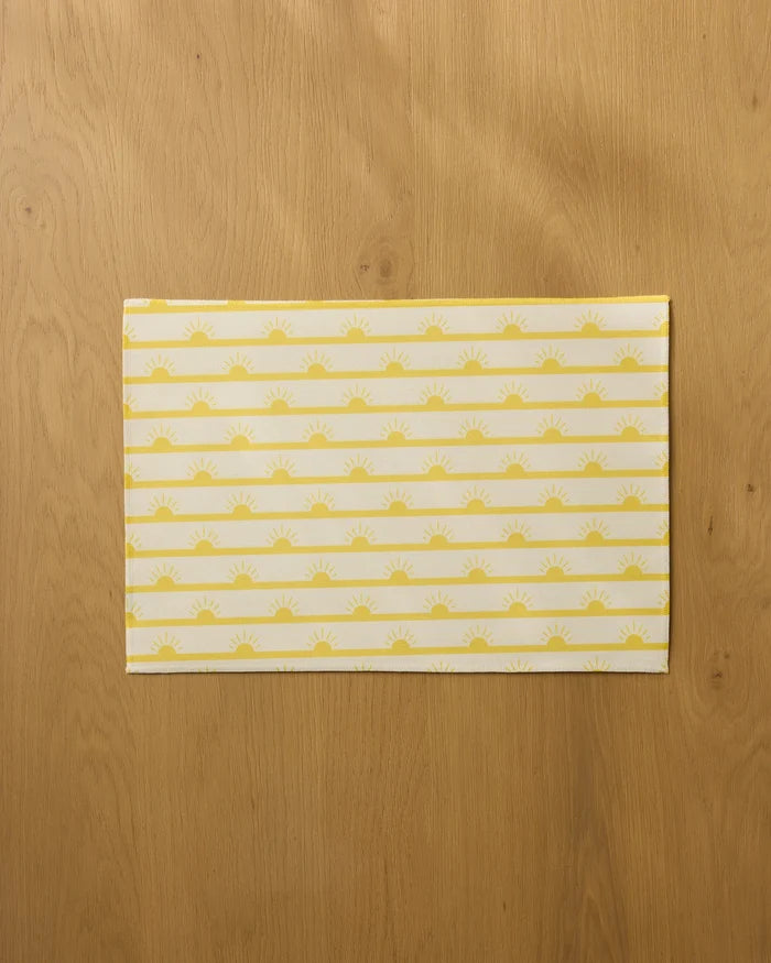 The Geometry Luminary placemat features yellow sunburst and stripe patterns on a light background, displayed on a wooden surface.