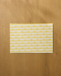 The Geometry Luminary placemat features yellow sunburst and stripe patterns on a light background, displayed on a wooden surface.