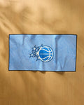 The NBALAB x Geometry Magic Grip, a blue NBA fitness towel with a basketball and star logo reminiscent of the Orlando Magic emblem, lies flat on a light wooden surface.