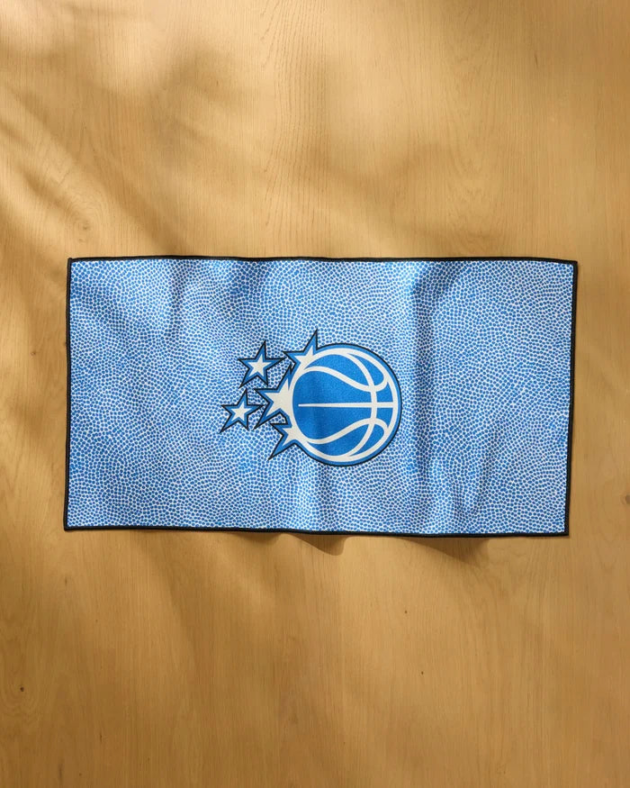 The NBALAB x Geometry Magic Grip, a blue NBA fitness towel with a basketball and star logo reminiscent of the Orlando Magic emblem, lies flat on a light wooden surface.