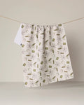 A white kitchen towel from Geometry, called “Martini Tiles,” featuring green and brown gift-themed designs and made from recycled materials, hangs on a string against a plain beige background.