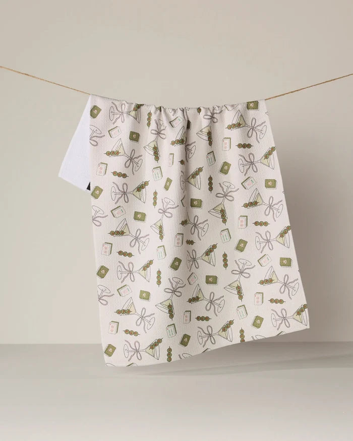 A white kitchen towel from Geometry, called “Martini Tiles,” featuring green and brown gift-themed designs and made from recycled materials, hangs on a string against a plain beige background.