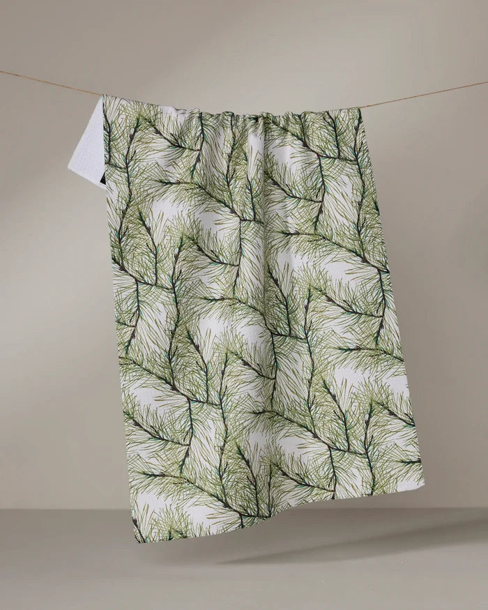 The Geometry Pinewell recycled kitchen towel, featuring a green pine needle pattern, hangs naturally on a line against a beige background, highlighting its bold botanical design.