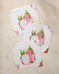Three white napkins from Geometry's Pink Dragon dishcloth set, featuring vibrant dragon fruit prints, are neatly arranged on a light beige marble surface.