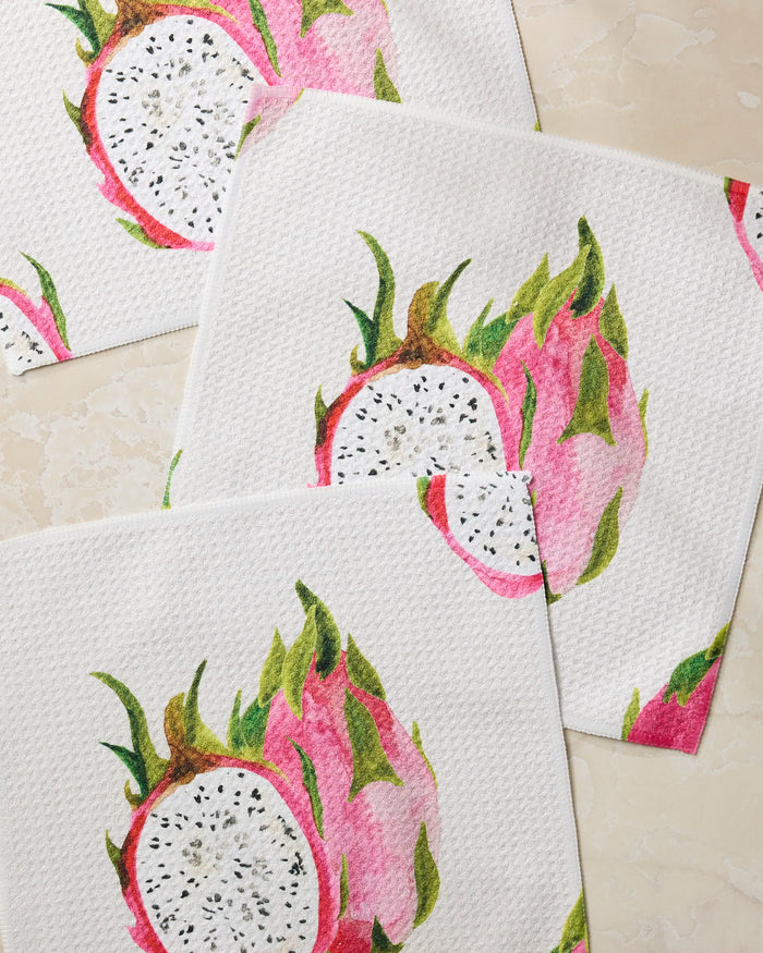Three Pink Dragon napkins by Geometry, featuring vibrant dragon fruit designs with pink and green skin, white flesh, and black seeds, are arranged overlapping on a light marble-textured surface.